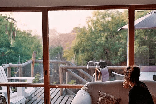 View from inside a living room through tall glass windows onto a wooded deck, with a woman sitting on a gray sofa